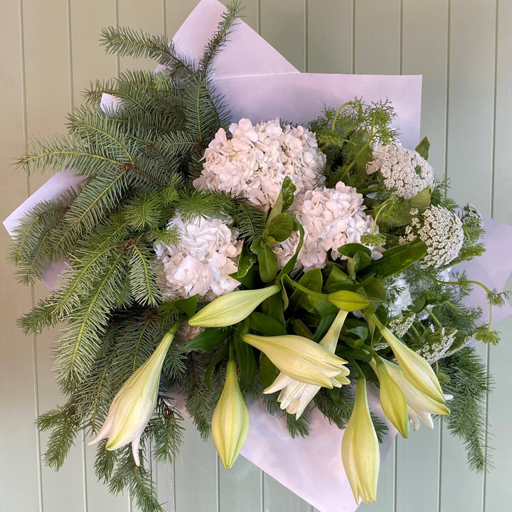 A Christmas-themed floral arrangement featuring lilies, hydrangeas, and other holiday foliage, presented against a green background.