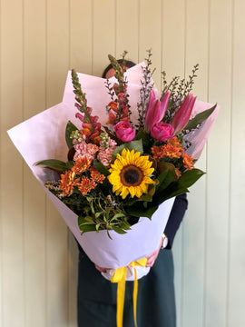 A person holds the COCKTAIL BUNCH (MODEST) by Smellies Flowers—a vibrant mix of Australian blooms, including sunflowers, pink lilies, and orange flowers wrapped in white paper—displayed against a light-paneled wall.