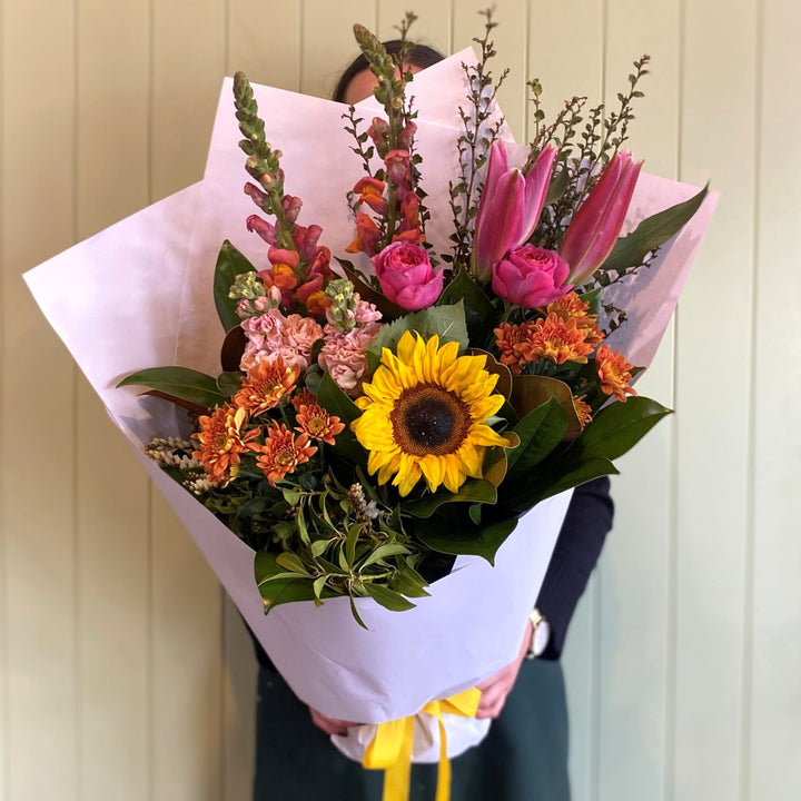 A person holds the COCKTAIL BUNCH (MODEST) by Smellies Flowers—a vibrant mix of Australian blooms, including sunflowers, pink lilies, and orange flowers wrapped in white paper—displayed against a light-paneled wall.