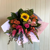 The COCKTAIL BUNCH (MODEST) by Smellies Flowers showcases seasonal sunflowers, pink tulips, roses, and orange blooms in white wrapping, set against a light wooden background—celebrating local beauty and sustainable floristry.