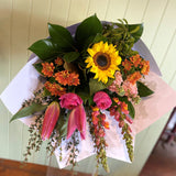 The COCKTAIL BUNCH (MODEST) by Smellies Flowers is a vibrant bouquet with sunflowers, pink lilies, roses, and snapdragons in lush greenery, wrapped sustainably in white paper—displayed against light wood décor.