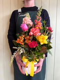 A person in a dark sweater and green apron holds the Smellies Flowers COCKTAIL FUSS POT—a white vase with yellow ribbon, filled with colorful Australian-grown flowers—against a light wooden background.