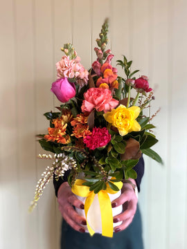 A person holds the COCKTAIL FUSS POT by Smellies Flowers—a vibrant mix of pink, yellow, and orange blooms with green foliage in a mini ceramic vase tied with a yellow and white ribbon. The background is softly blurred.