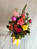 The COCKTAIL FUSS POT by Smellies Flowers is a vibrant mix of pink, red, orange, and yellow Australian blooms in a white vase with a yellow ribbon, set against a white paneled background.