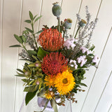Bouquet of flowers with red and orange proteas, yellow daisies, and greenery in a clear vase against a white wooden background.