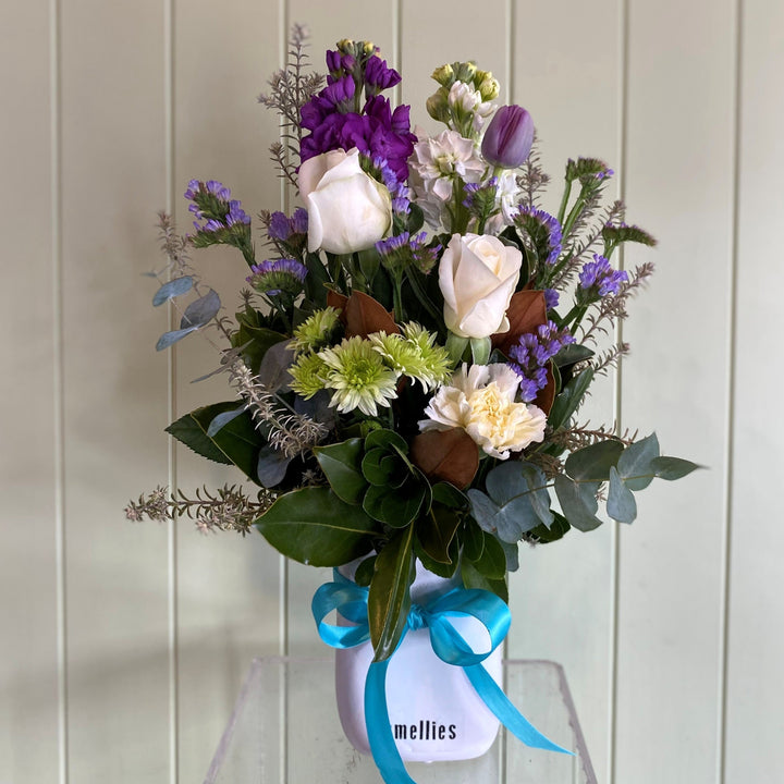 Small white ceramic pot of purple and mauve flowers with a blue ribbon on a white background