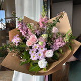 Bouquet of soft, pastel flowers in pinks and mauve, wrapped in brown paper.