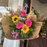 Bright bouquet of flowers with pink roses, sunflowers, and greenery in a brown paper wrap.