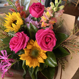 Bright bouquet of flowers with hot pink roses, sunflowers, and other assorted flowers in brown paper.