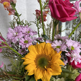 Arrangement of flowers including a yellow sunflower, hot pink and purple flowers, and green foliage.