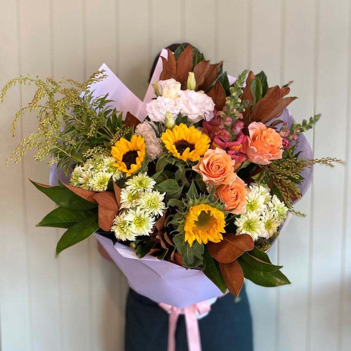 Person holding a large flower bouquet of roses, sunflowers, chrysanthemums, lisianthus and snapdragons with foliage wrapped in white paper