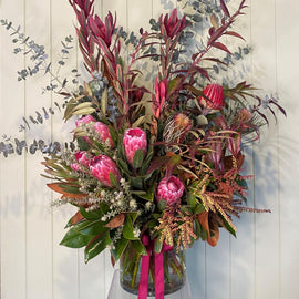 A vibrant floral arrangement in a clear glass vase filled with water, showcasing large native flowers like pink protea, magnolia leaves, and assorted greenery. It is tied with a burgundy ribbon and set against a pale green paneled background