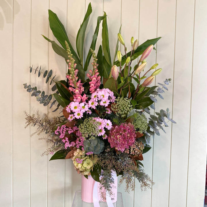 A large, elegantly arranged bouquet of flowers in a pale pink vase, perfect for welcoming a baby girl. The bouquet features pink lilies, snapdragons, assorted pink and purple flowers, and green foliage. Set against light-colored wooden paneling, this pretty arrangement is available for delivery in Geelong.