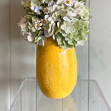 Yellow vase with flowers on a clear stand against a white paneled wall