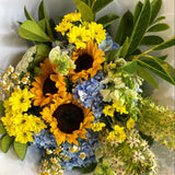 Bouquet of sunflowers, yellow and blue flowers, and greenery on a white paper background.
