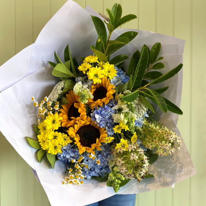 Bouquet of flowers wrapped in white paper held by a person wearing blue jeans against a light green wall.