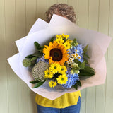 Person holding a bouquet of sunflowers and other flowers against a light green wall.