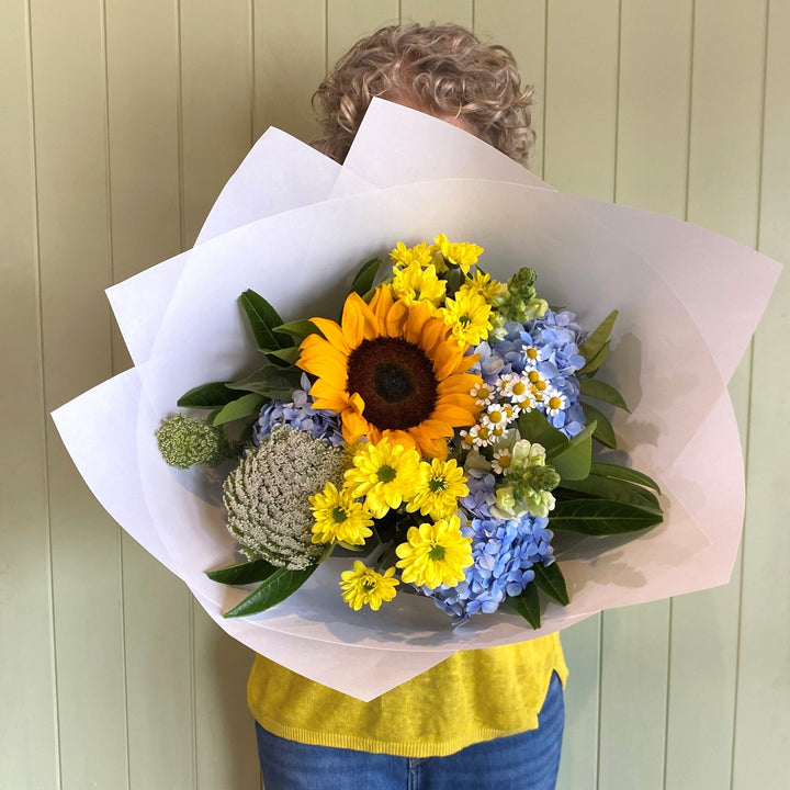 Person holding a bouquet of sunflowers and other flowers against a light green wall.