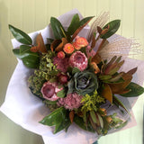 A eucalyptus and native bouquet featuring a mix of proteas, kale, leucadendrons, strawflowers, sedum, hydrangeas, and miscanthus grass with foliage, wrapped in white paper.