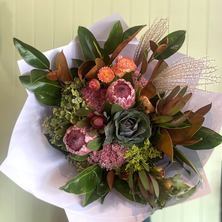 A eucalyptus and native bouquet featuring a mix of proteas, kale, leucadendrons, strawflowers, sedum, hydrangeas, and miscanthus grass with foliage, wrapped in white paper.