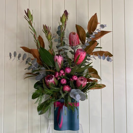 A floral arrangement featuring various pink proteas, eucalyptus leaves, and green foliage, displayed in a teal vase with a pink ribbon. The vase sits on a clear stand against a light-colored, vertical-paneled background. This bouquet of native flowers exudes an earthy charm