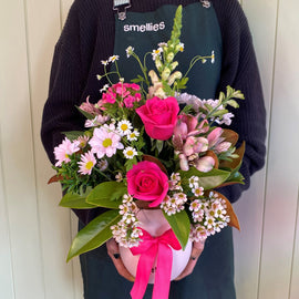 A person wearing a dark sweater and green apron holds a vibrant bouquet of pretty flowers, including pink roses, white daisies, and various greenery, all tied with a pink ribbon.