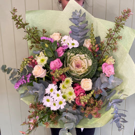 A person holds a large bouquet featuring seasonal flowers, including pink roses, white daisies, and other green and pink blooms. The bouquet is wrapped in light green floral paper, standing against a light green paneled background. Perfect for Geelong flower delivery enthusiasts seeking fresh elegance.