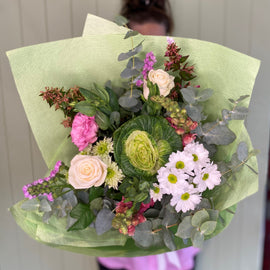 A person holds a large bouquet featuring seasonal flowers, including pink roses, white daisies, and other green and pink blooms. The bouquet is wrapped in light green floral paper, standing against a light green paneled background. Perfect for Geelong flower delivery enthusiasts seeking fresh elegance.