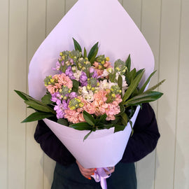 Person in a dark outfit holding a large bouquet of pastel-colored cut flowers, including pink, purple, and white blossoms, wrapped in white paper with green leaves. The background features vertical wooden panels in a light color. The person's face is obscured by the flowers. Perfect for Geelong flower delivery!