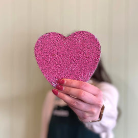 Hand holding a pink glittery heart-shaped object against a neutral background
