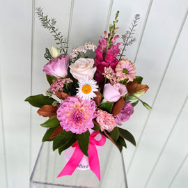 A floral arrangement in a white cylindrical vase with the text "Lillies". The bouquet includes pink roses, carnations, dahlias, white daisies, and assorted greenery, accented with a hot pink ribbon tied around the vase. These pretty flowers are set against a light green, paneled backdrop.