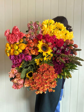 A person holds Smellies Flowers' COCKTAIL POSIE OF THE DAY—a vibrant bouquet—in front of a light-colored, vertical wooden wall.