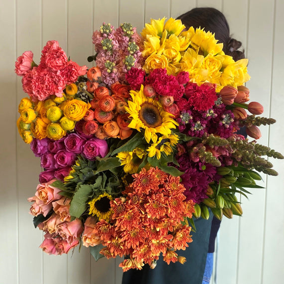 A person holds Smellies Flowers' COCKTAIL POSIE OF THE DAY—a vibrant bouquet—in front of a light-colored, vertical wooden wall.