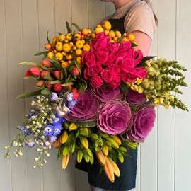 A person holding the RAINBOW POSIE OF THE DAY from Smellies, a large and colorful bouquet featuring pink roses, orange and yellow flowers, blue delphiniums, purple ornamental cabbages, and various green leaves sourced from Australian flower farms. The premium blooms are arranged vibrantly against a plain background.