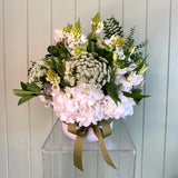 White pot of flowers with white hydrangeas and other white flowers on a clear stand against a light wooden panel background