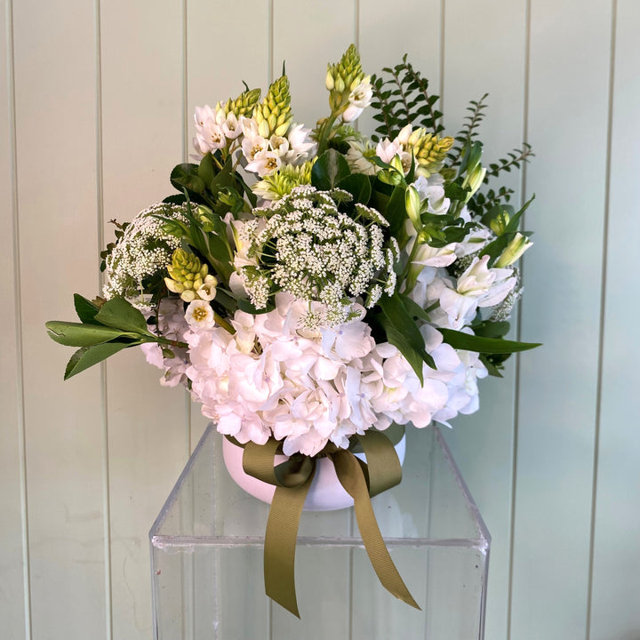White pot of flowers with white hydrangeas and other white flowers on a clear stand against a light wooden panel background