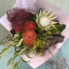 A floral arrangement featuring a variety of native flowers including red and green proteas, white and orange lilies, and green foliage, wrapped in a white paper.