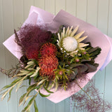 A floral arrangement featuring a variety of native flowers including red and green proteas, white and orange lilies, and green foliage, wrapped in a white paper.