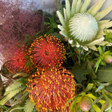 A floral arrangement featuring a variety of native flowers including red and green proteas, white and orange lilies, and green foliage, wrapped in a white paper.