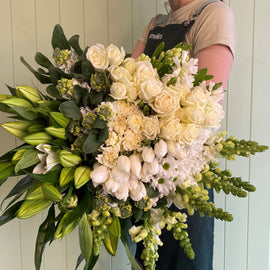 A person holds a large bouquet of predominantly white flowers, including roses, lilies, tulips, and snapdragons. The elegant arrangement is lush with green foliage and cascades beautifully. Clad in a dark green apron, the person presents this classic floral masterpiece with grace.