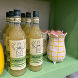 Bottles of Margarita juice on a green shelf with a decorative pitcher.