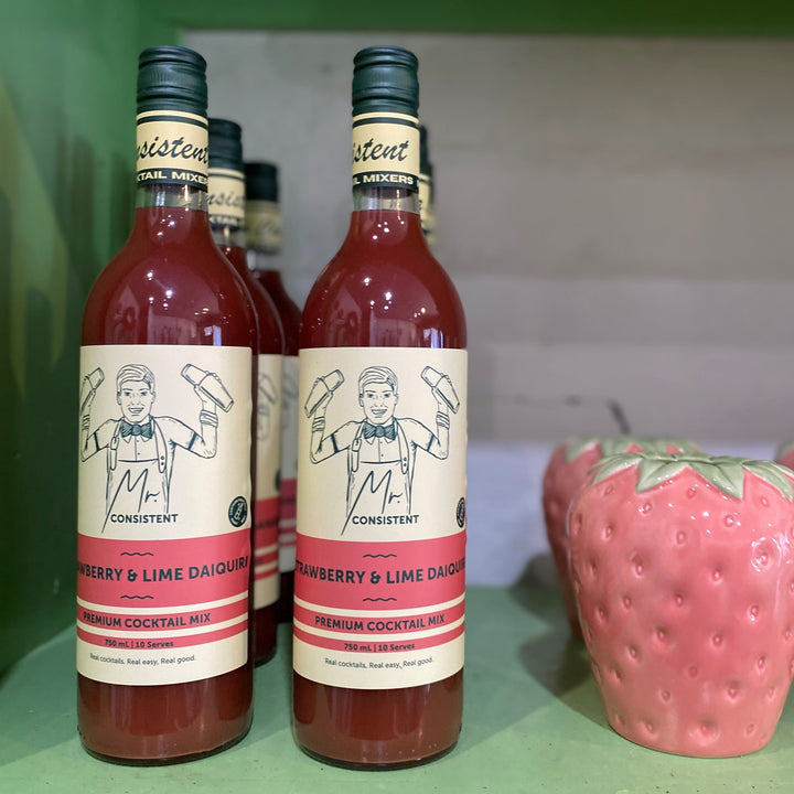 Bottles of berry and lime daiquiri mix with strawberry-shaped container on a green surface.